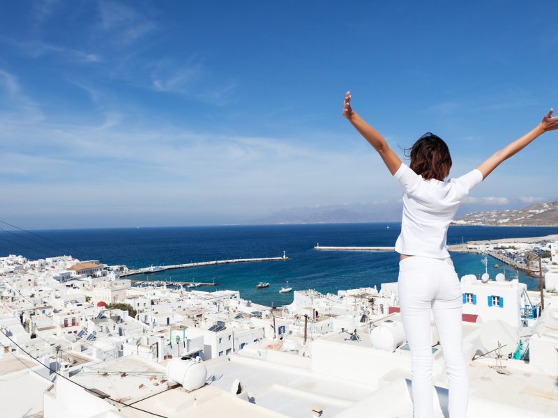 mykonosRear view of young woman with arms outstretched, looking at the cityscape of Mykonos island, Greece.